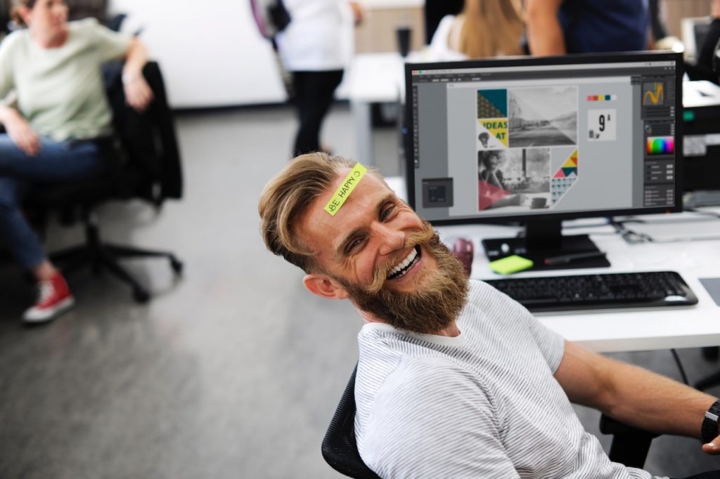 Un homme sourit à son bureau avec un post-it sur le front.