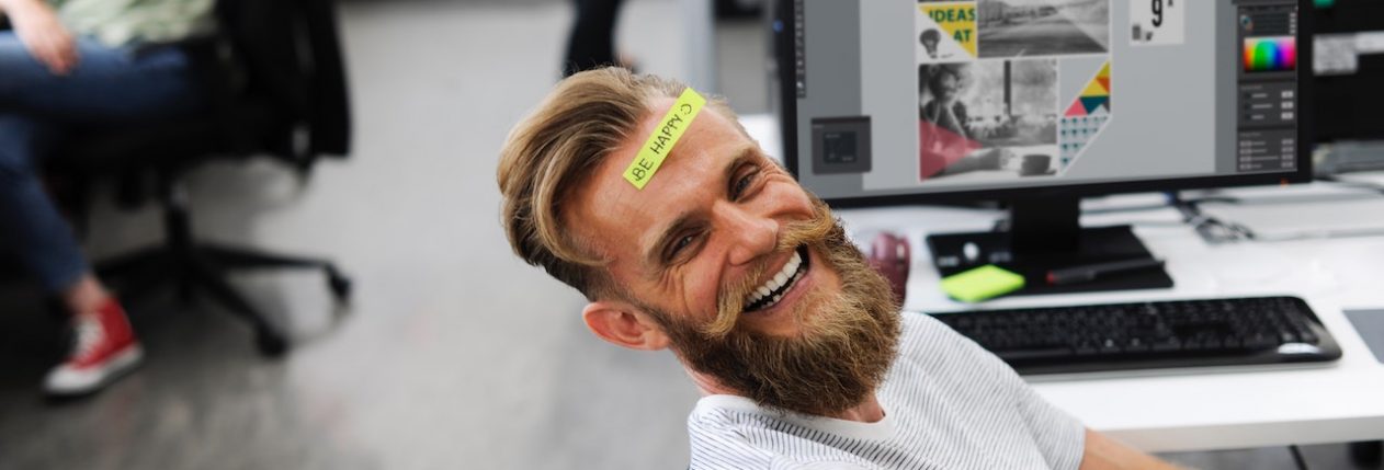 Un homme sourit à son bureau avec un post-it sur le front.