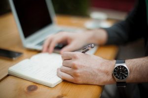 Un homme ecrit sur un bloc-note à côté d'un ordinateur.