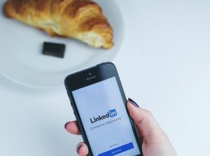 Main de femme tenant un smartphone sur la page de LinkedIn. À côté il y a une assiette avec un croissant et du chocolat dessus.