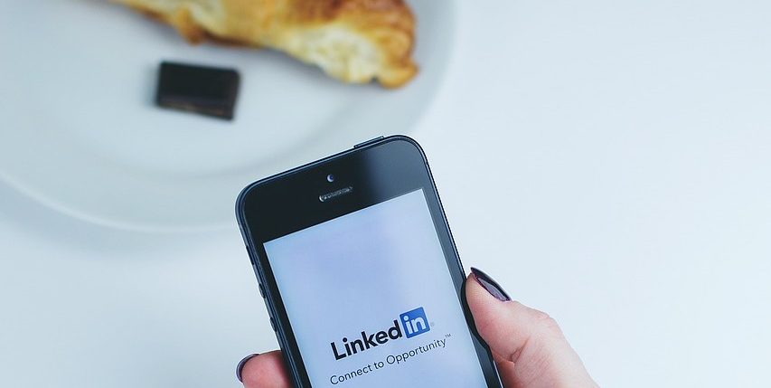 Main de femme tenant un smartphone sur la page de LinkedIn. À côté il y a une assiette avec un croissant et du chocolat dessus.