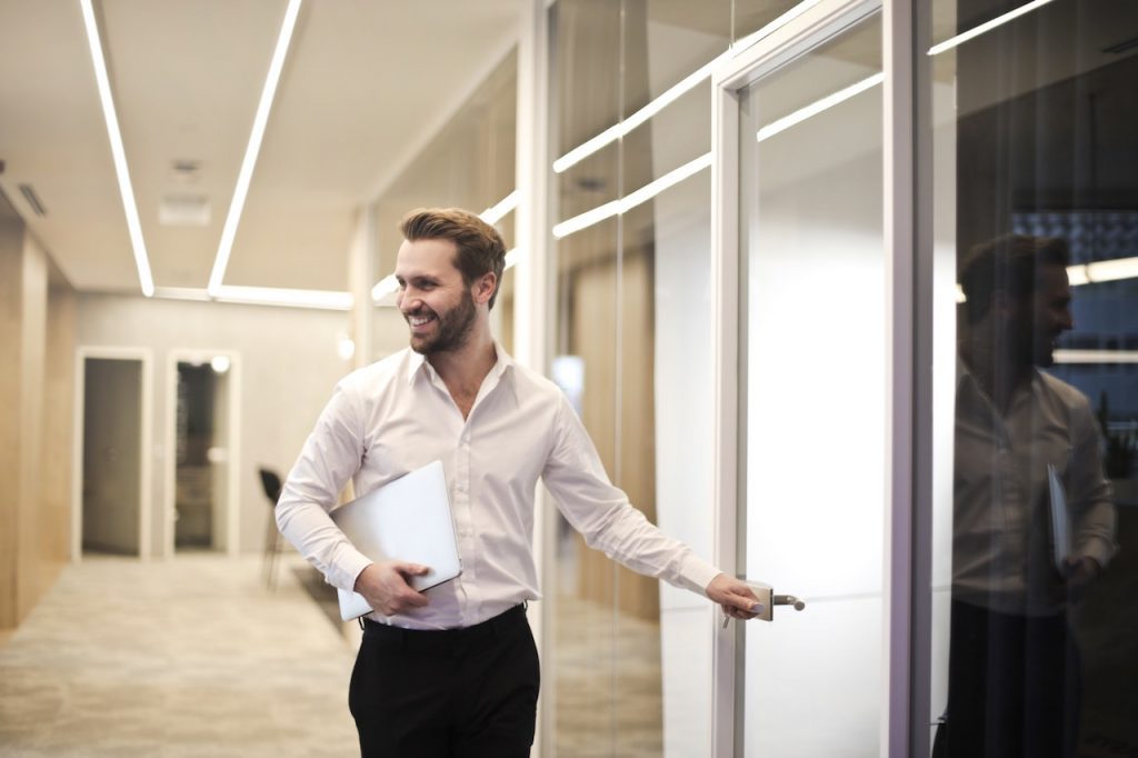 Un homme en chemise blanche sourit. Il est dans un couloir et il s'apprête à ouvrir une porte.