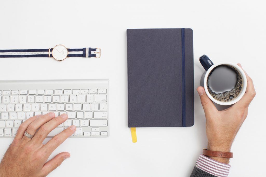 Un bureau blanc avec un clavier, une montre, un bloc-note et une main qui tient une tasse de café.