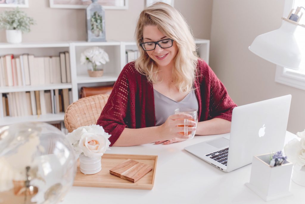 Une femme souriante est assise à une table devant son ordinateur avec une tasse de thé à la main.