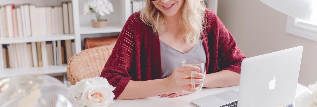 Une femme souriante est assise à une table devant son ordinateur avec une tasse de thé à la main.