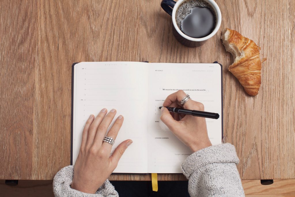 Une femme écrit dans son journal à côté d'un café et d'un croissant.