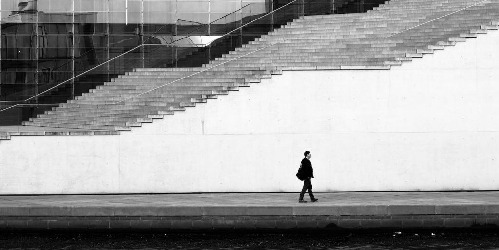 Un homme marche devant un bâtiment avec un grand escalier, à l'entrée. La photo est en noir et blanc.