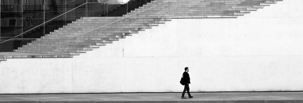 Un homme marche devant un bâtiment avec un grand escalier, à l'entrée. La photo est en noir et blanc.