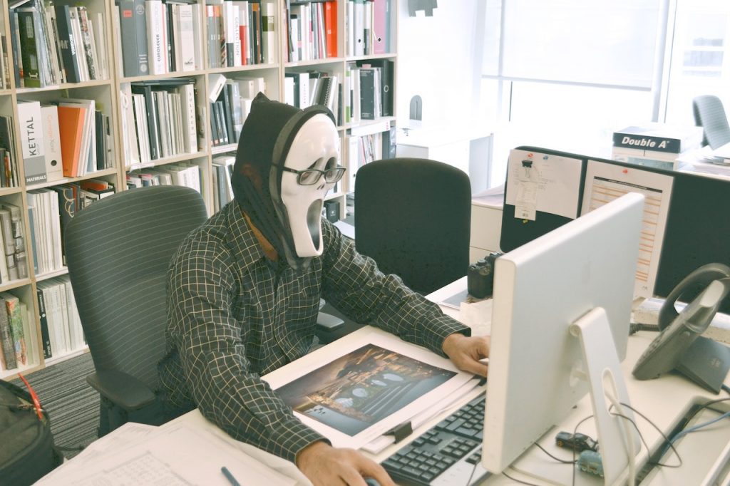 Un homme assis à un bureau devant un ordinateur. Il porte le masque de Scream.