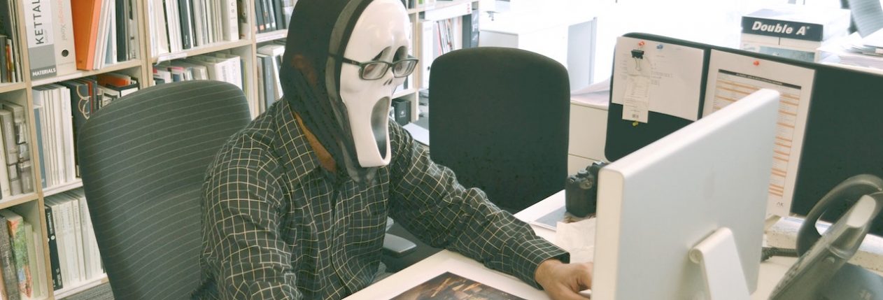 Un homme assis à un bureau devant un ordinateur. Il porte le masque de Scream.