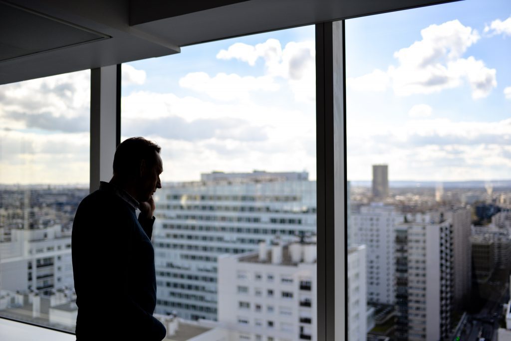 Un homme de dos devant une fenêtre donnant sur une ville avec des gratte-ciels. Il est au téléphone.