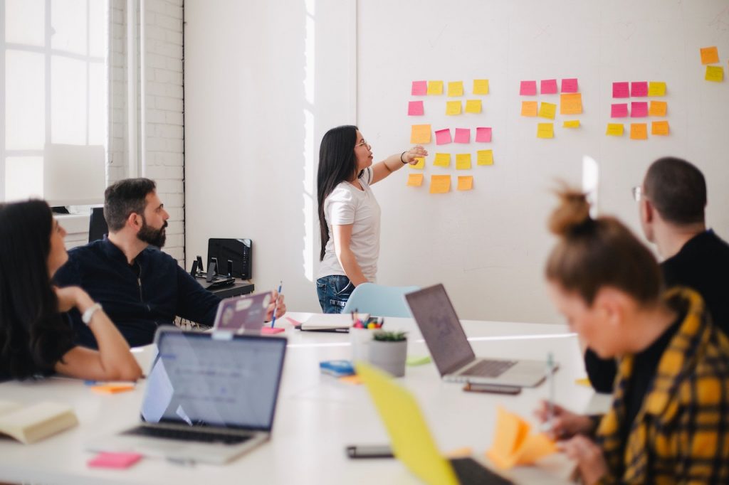 Un groupe de travail autour d'une table. Une femme se trouve devant un tableau blanc avec plusieurs post-it, devant le groupe.