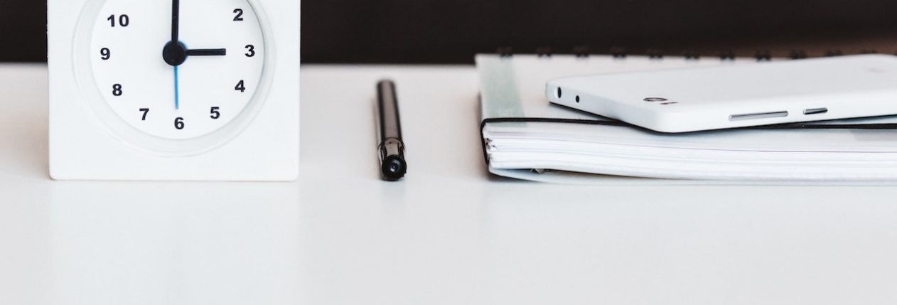 Une horloge blanche, posée sur une table à côté d'un carnet, d'un stylo et d'un téléphone.