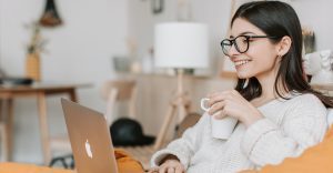 Une femme assie face à un ordinateur. Elle tient un mug blanc et elle sourit.