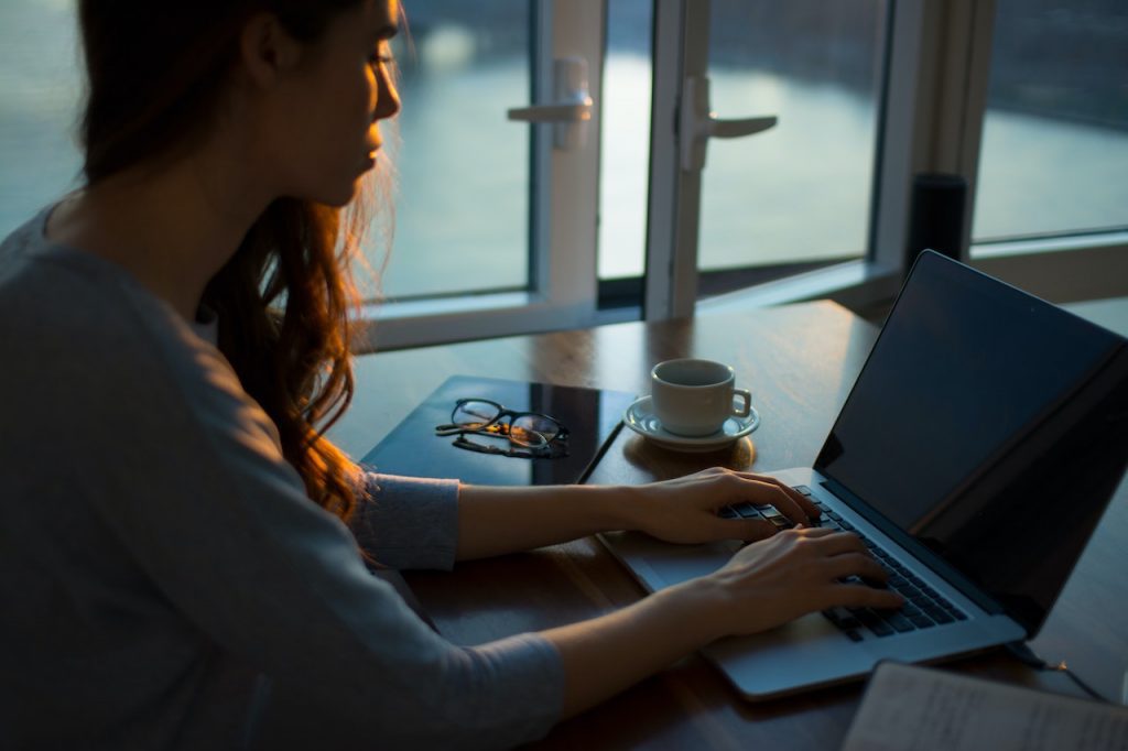 Une femme assise à une table près d'une fenêtre. Elle tape sur un ordinateur portable. À côté d'elle, il y a un mug, un carnet et des lunettes de vue.