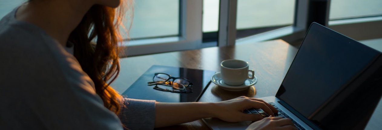 Une femme assise à une table près d'une fenêtre. Elle tape sur un ordinateur portable. À côté d'elle, il y a un mug, un carnet et des lunettes de vue.