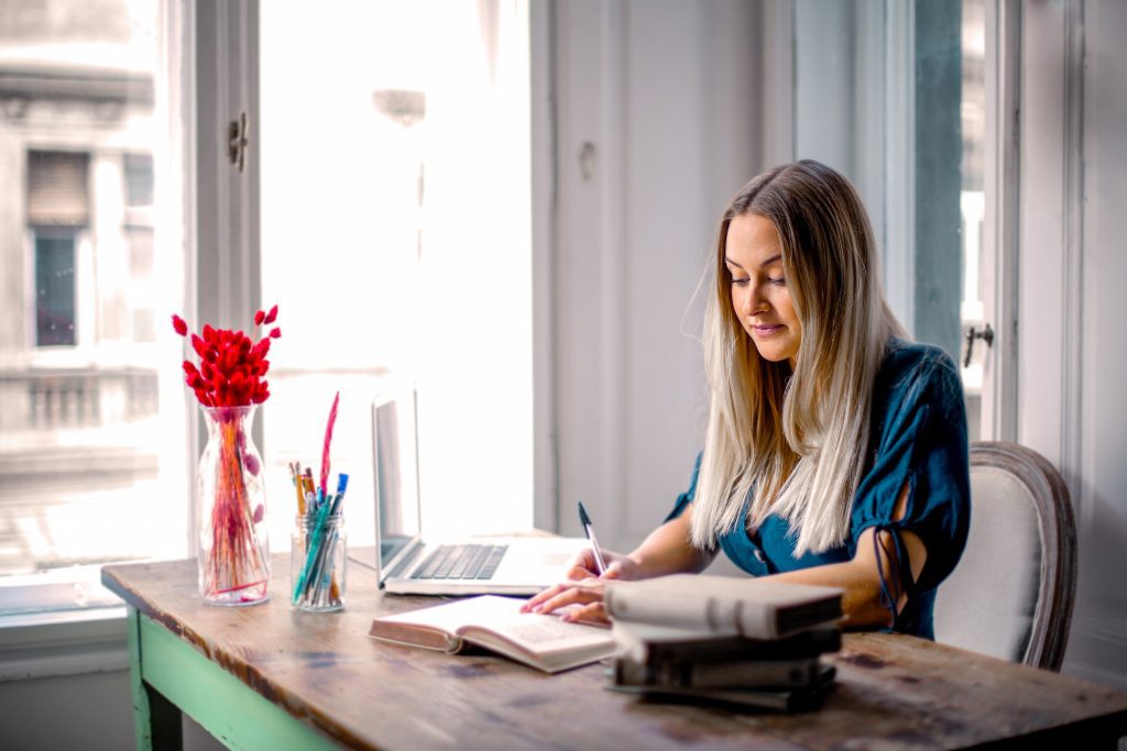 Une femme blonde avec une chemise bleue est assise à son bureau devant une fenêtre. Elle feuillette un carnet.