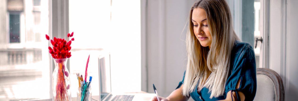 Une femme blonde avec une chemise bleue est assise à son bureau devant une fenêtre. Elle feuillette un carnet.
