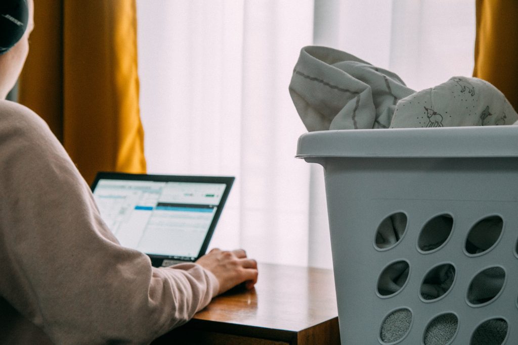 Une personne de dos est assis à une table devant un ordinateur portable. On voit son écran. Au premier plan, on voit un panier à linge rempli à ras bord.