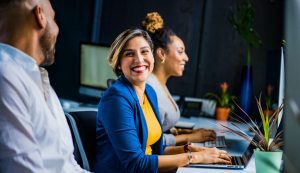 Trois personnes assises l'une à côté de l'autre, face à des ordinateurs. La personne au milieu est une femme et elle sourit à la personne à côté d'elle.