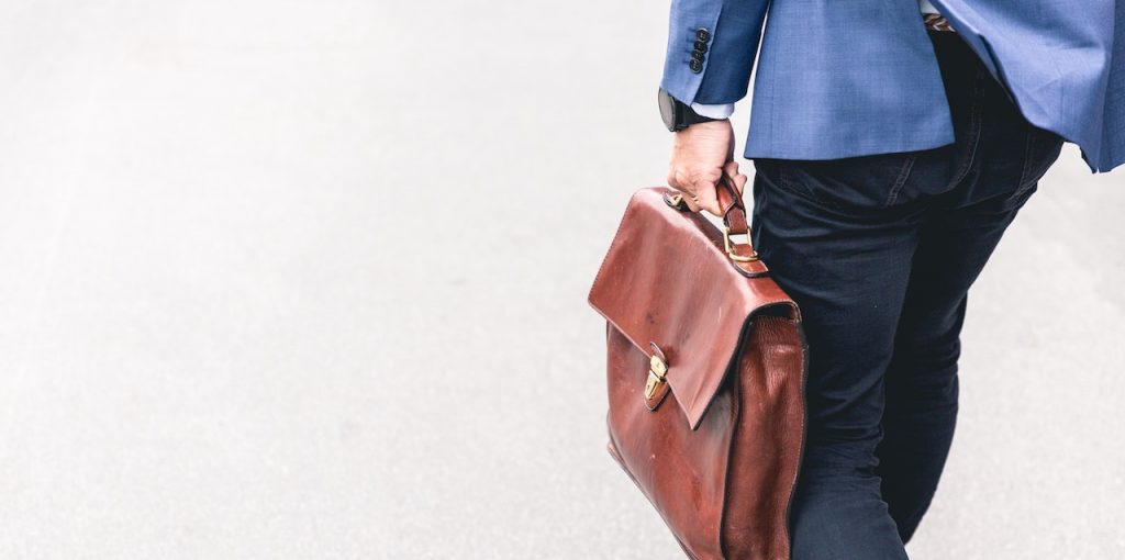 On voit un homme de dos en train de marcher. Il porte un pantalon noir, une veste de costume bleu et il tient à la main une sacoche en cuir marron.