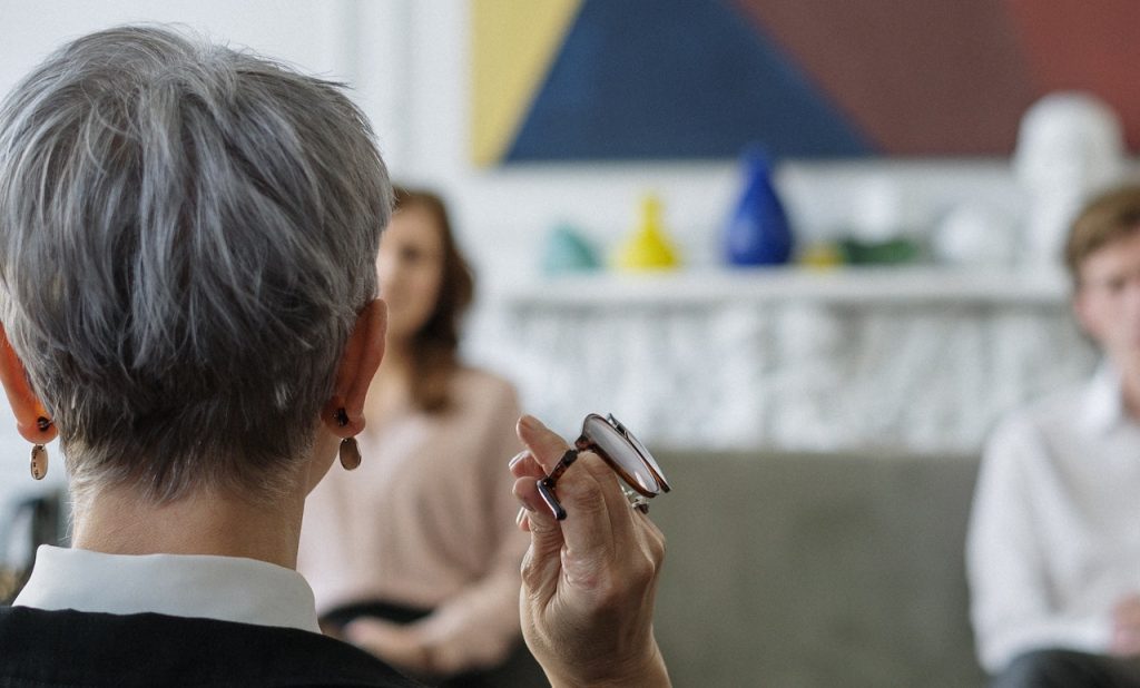 On voit une femme au cheveux courts et gris de dos. Elle tient des lunettes et elle fait face à deux personnes assises un peu plus loin devant elle. Ces personnes sont floues sur la photo.