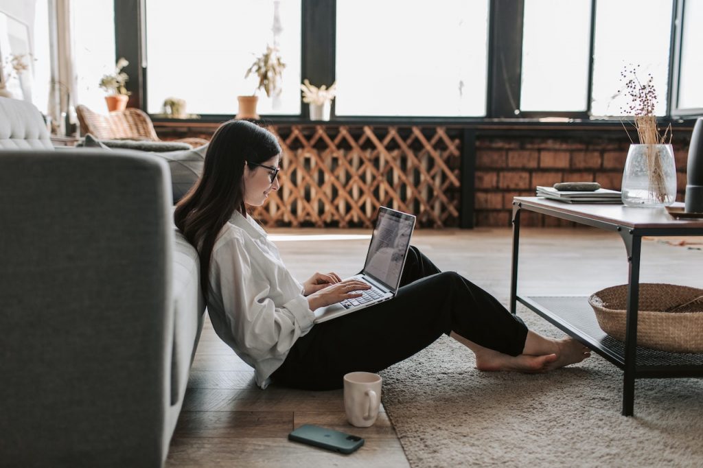 Une femme assise par terre, adossée sur un canapé. Elle a son ordinateur sur les genou et son téléphone et un mug par terre à côtté d'elle. Elle sourit.