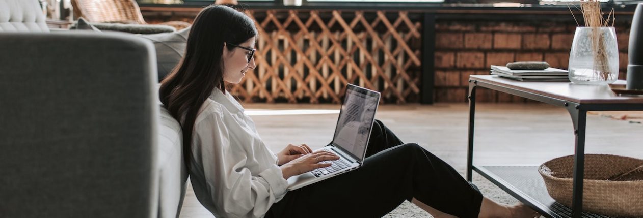Une femme assise par terre, adossée sur un canapé. Elle a son ordinateur sur les genou et son téléphone et un mug par terre à côtté d'elle. Elle sourit.