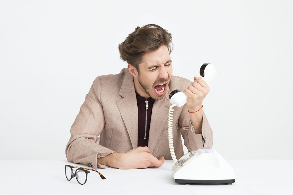 Un homme jeune en costume clair est assis à une table blache. Le fond derrière lui est blanc aussi. Il y a un vieux téléphone blanc sur la table et une paire de lunettes de vues. L'homme tient le récepteur du téléphone à la main et il semble crier sur la personne de l'autre côté du téléphone.