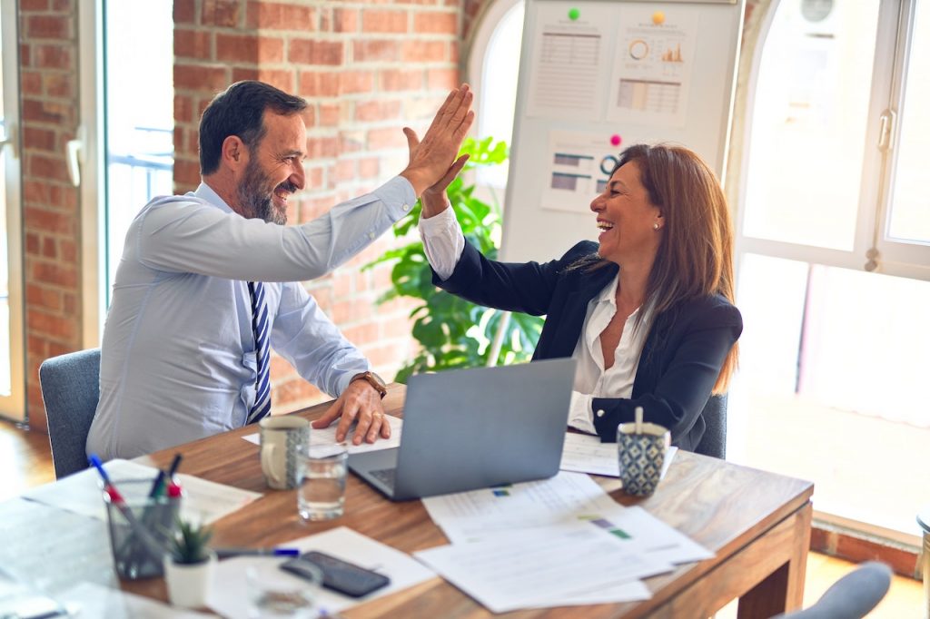 Un homme et une femme sont assis à une table. Ils rigolent et se tapent dans la main en signe de victoire. Sur la table il y a des dossiers et un ordinateur portable.