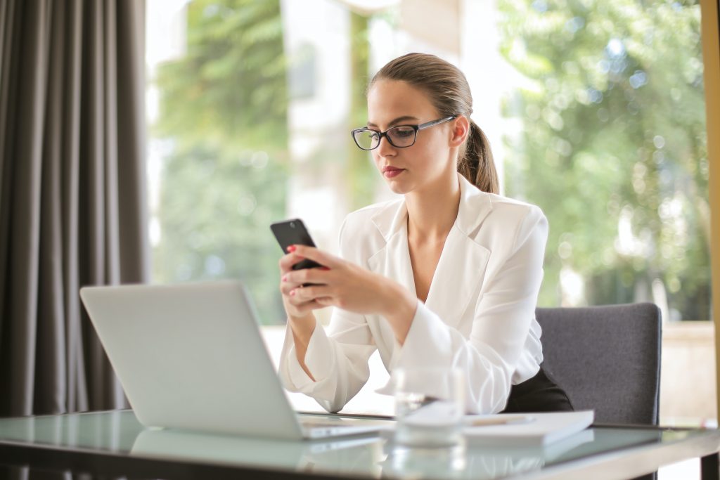 Une femme assise devant un bureau face à un ordinateur portable ouvert. Le fond est une fenêtre et on voit la nature à l'extérieur. Elle porte des lunettes, une queue de cheval et une chemise blance. Elle tient un téléphone portable à hauteur de ses yeux et elle semble envoyer un SMS.