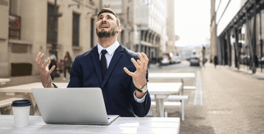 Un homme en costume assis à une terrasse face à son ordinateur portable. Il regarde le ciel avec un air désespéré et ses mains lui donnent l'air d'être frustré