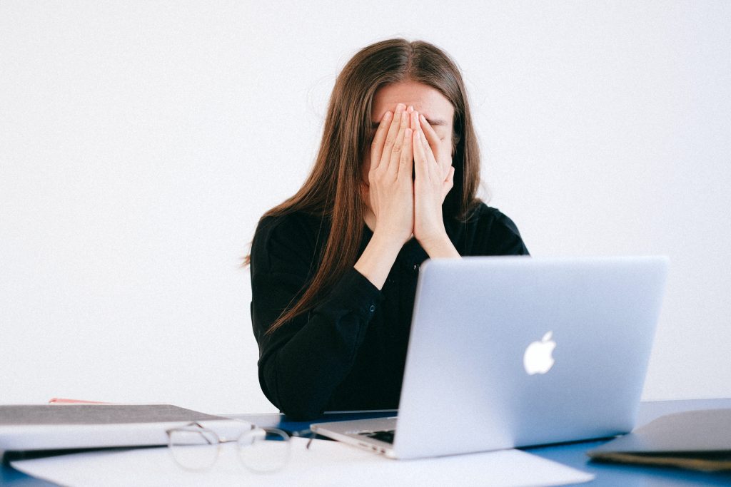 Une femme assise à une table devant un ordinateur portable ouvert. Elle se cache le visage avec les mains ; elle semble fatiguée ou stressée.