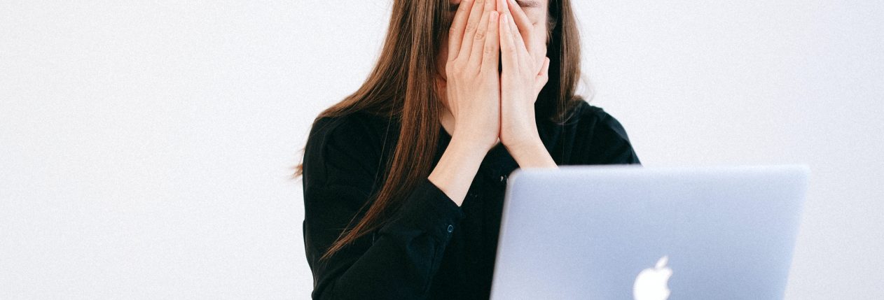 Une femme assise à une table devant un ordinateur portable ouvert. Elle se cache le visage avec les mains ; elle semble fatiguée ou stressée.