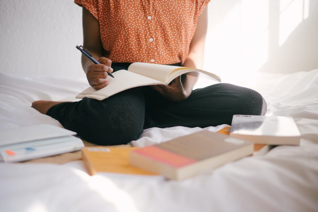 Une femme assise sur un lit avec plein de livres autour d'elle. Elle a un livre ouvert devant elle et elle tient un crayon.