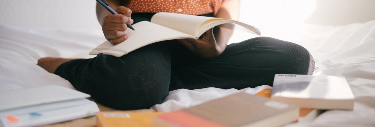 Une femme assise sur un lit avec plein de livres autour d'elle. Elle a un livre ouvert devant elle et elle tient un crayon.
