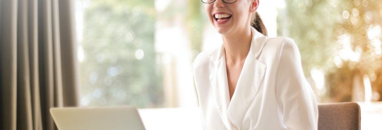 Une femme assise devant son ordinateur semble rire avec quelqu'un qui se trouve hors de la photo.
