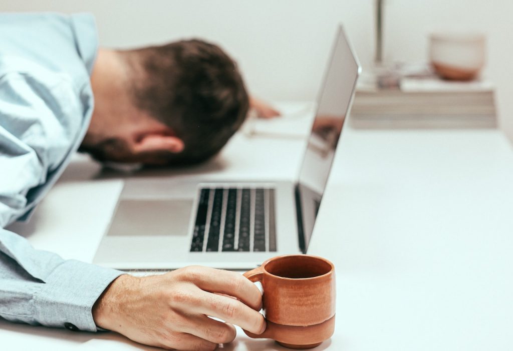 Un homme avec la tête posé sur une table à côté de son ordinateur. Il porte une tasse de café et a l'air de dormir.