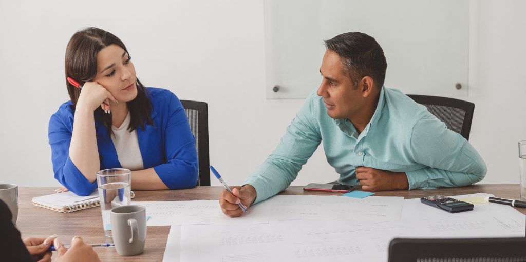 Un homme et une femme assis côte à côté à une table pendant une réunion de travail. La femme regrde l'homme, car il parle.