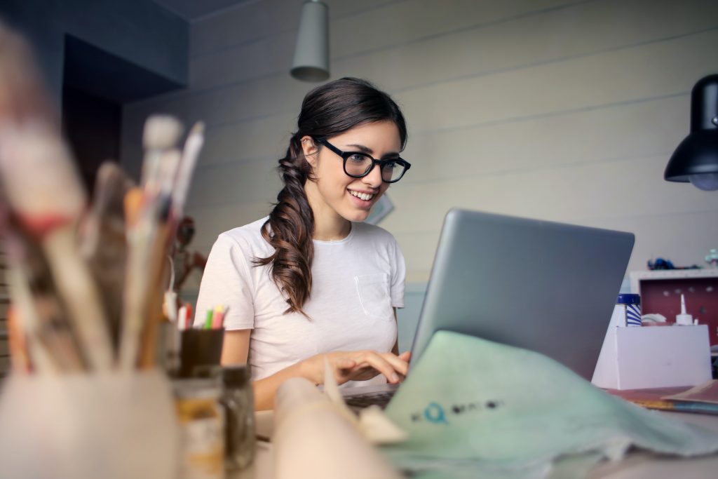 Une femme qui sourit devant un ordinateur. Il y a des pinceaux et de la peinture sur son bureau.