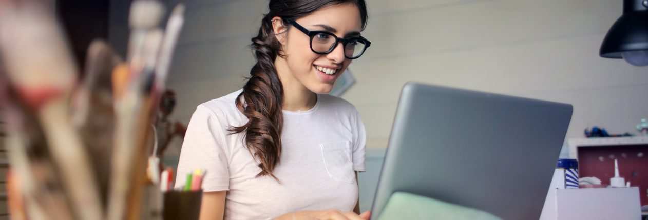 Une femme qui sourit devant un ordinateur. Il y a des pinceaux et de la peinture sur son bureau.