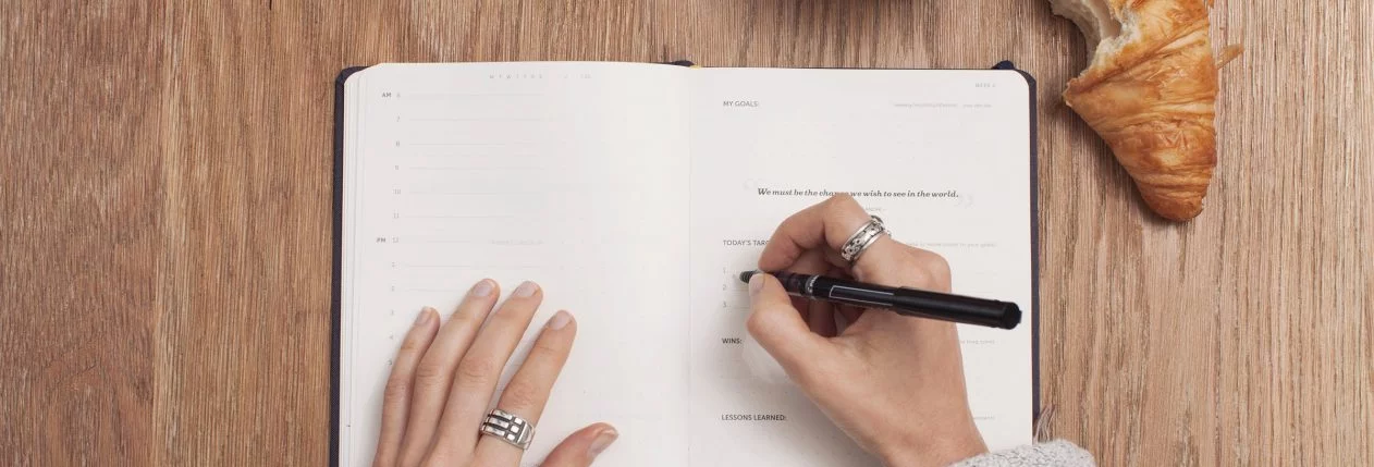 Une femme écrit dans son journal à côté d'un café et d'un croissant.