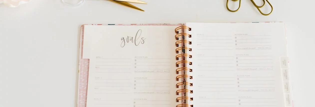 Un agenda ouvert sur une table blanche avec des fournitures dorées autour.