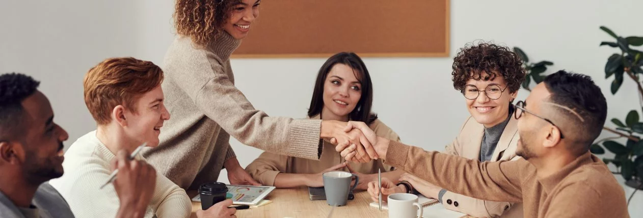 Un groupe de personnes assises autour d'une table. Une femme est debout et serre la main à un homme assis en face d'elle.