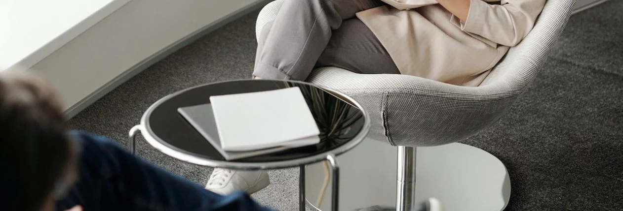 Deux personnes assises l'une en face de l'autre avec une table basse entre eux. L'une d'elles est assise avec les jambes croisées et des papiers à la main. L'autre personne est assise un peu plus nonchalamment et est en train de parler.