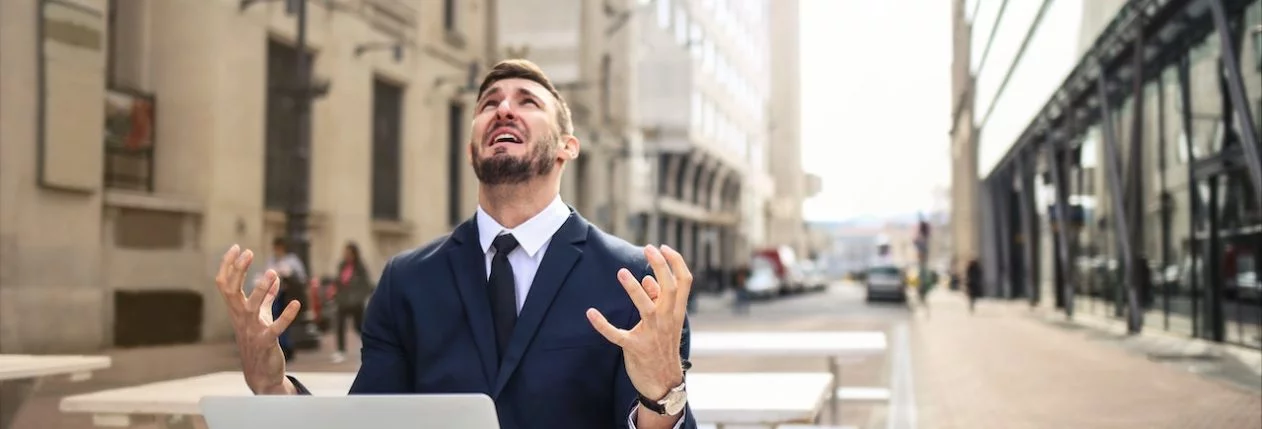 Un homme assis en terrasse avec son ordinateur. Il lève les bras au ciel et il a l'air désespéré.