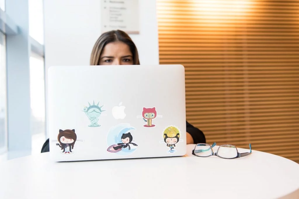 Une femme sur un ordinateur portable. On ne voit que ses yeux qui dépasse au-dessus de l'écran et on peut voir qu'elle a des stickers rigolos collés sur son ordinateur.